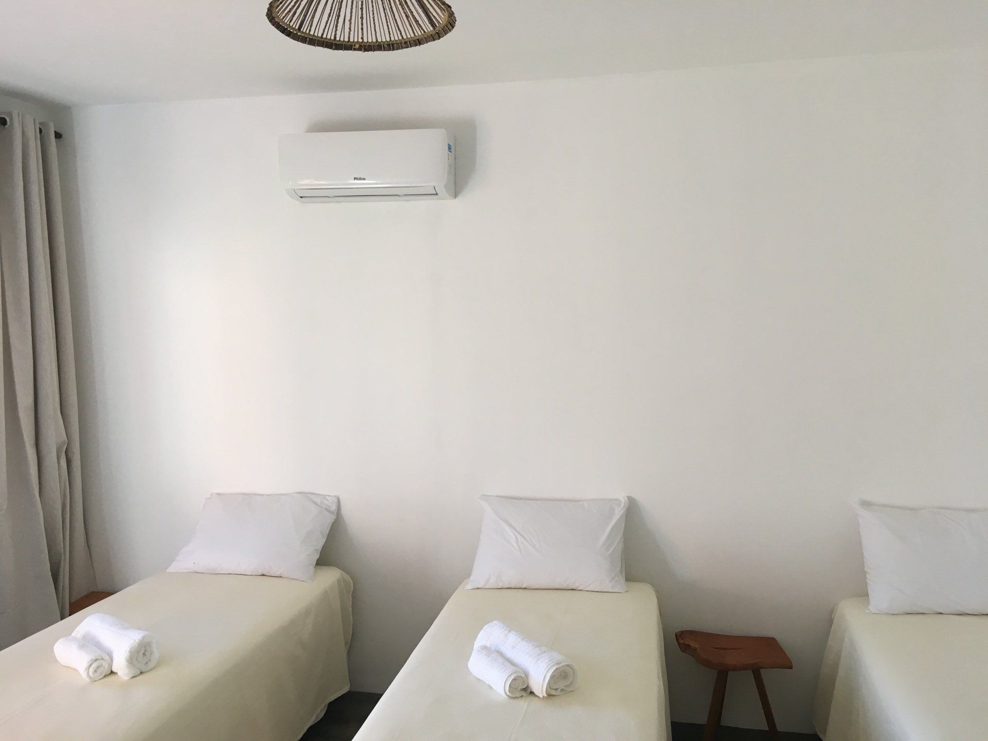 Três camas de solteiro em um quarto branco com ar condicionado e um pequeno banquinho de madeira.