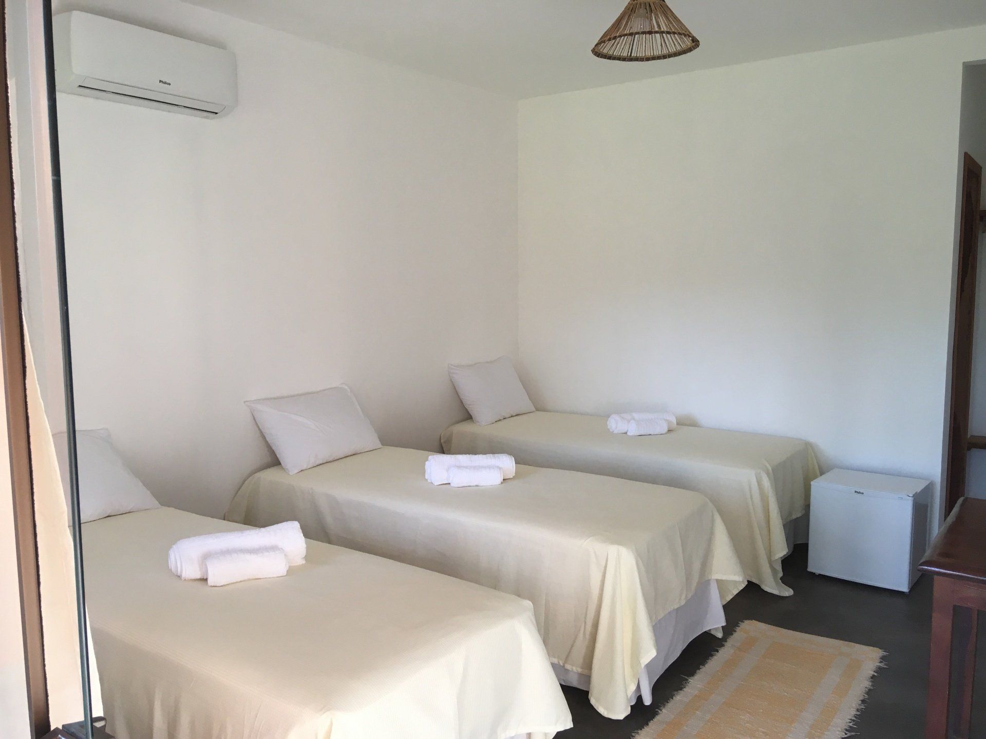 Três camas de solteiro em um quarto com paredes brancas, ar condicionado, um tapete e uma pequena geladeira.