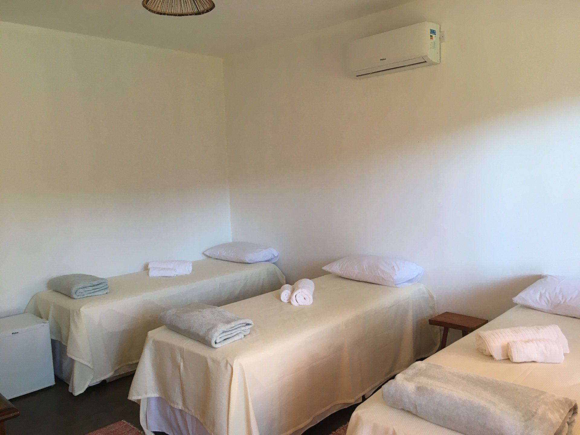 Três mesas de massagem em uma sala com paredes brancas, cada uma com toalhas e travesseiros. Frigobar e ar-condicionado visíveis.