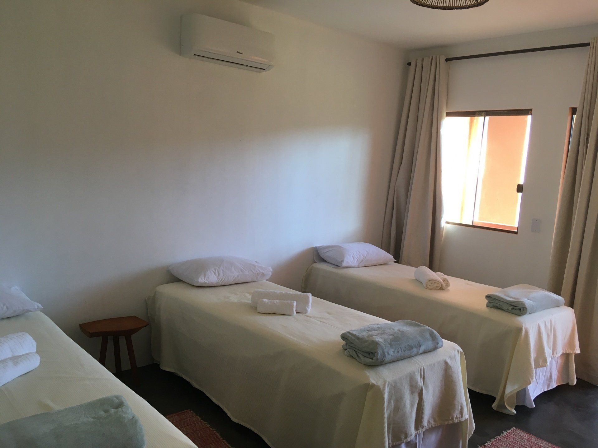 Três mesas de massagem em uma sala iluminada, cada uma com toalhas. Paredes brancas, cortinas e uma pequena mesa lateral.