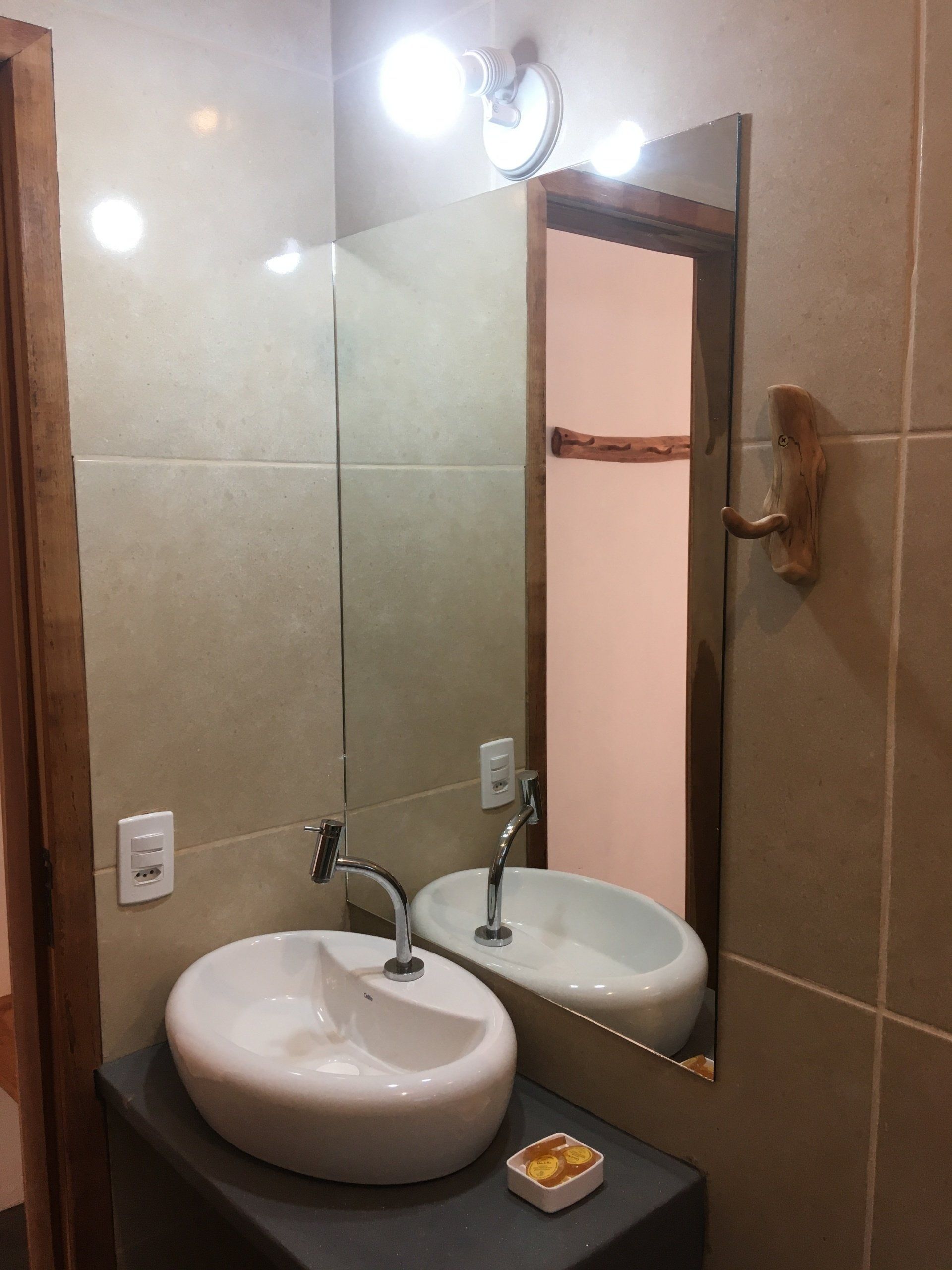 Banheiro com pia branca, espelho e luminária de parede, refletindo uma porta.