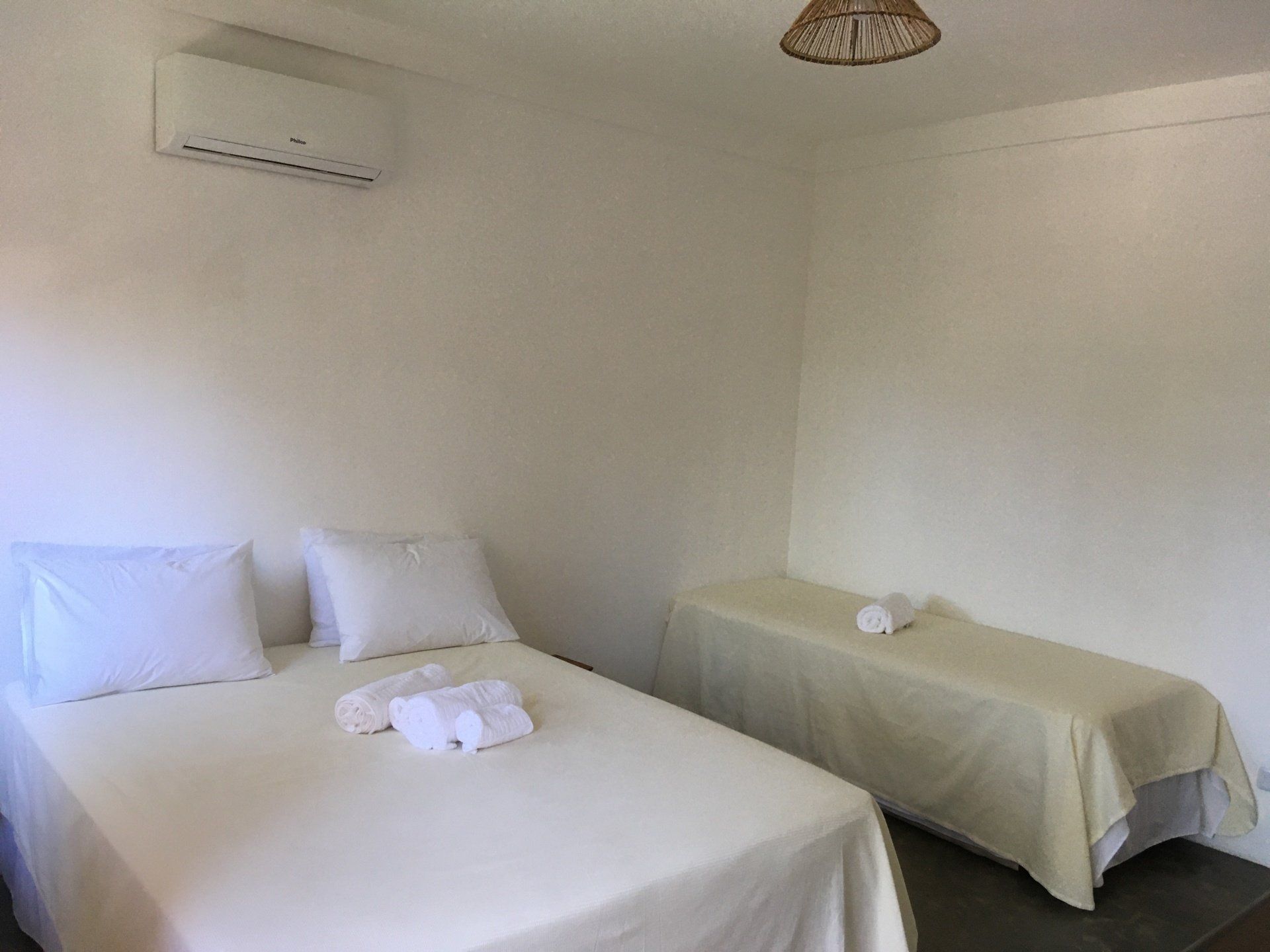 Um quarto com paredes brancas e duas camas, uma de casal e uma de solteiro, cobertas com roupa de cama branca.