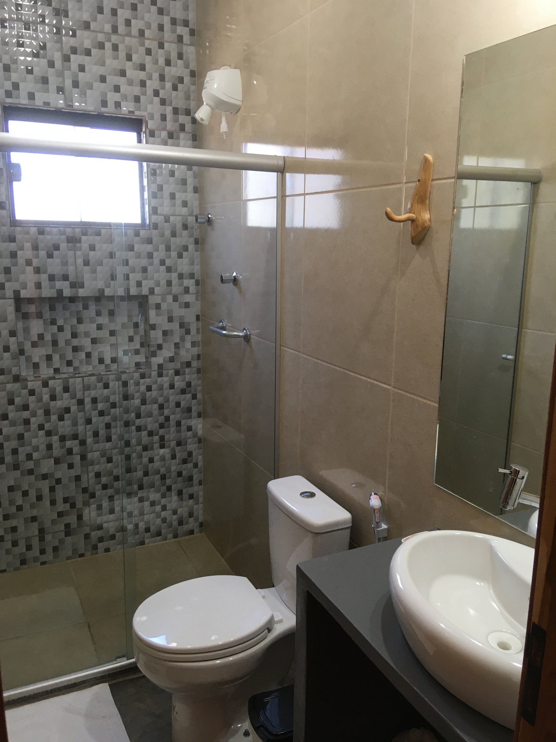 Banheiro com chuveiro, vaso sanitário, pia e espelho revestidos em azulejo. Esquema de cores bege e cinza.