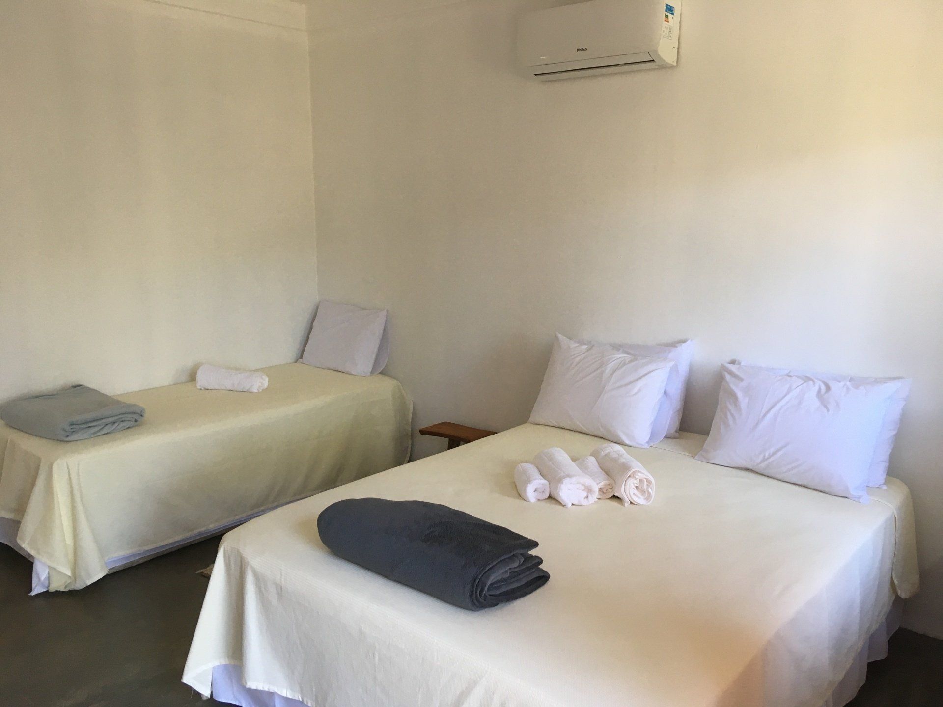 Duas camas em um quarto com paredes brancas, uma de solteiro e uma de casal, com roupa de cama e toalhas brancas.