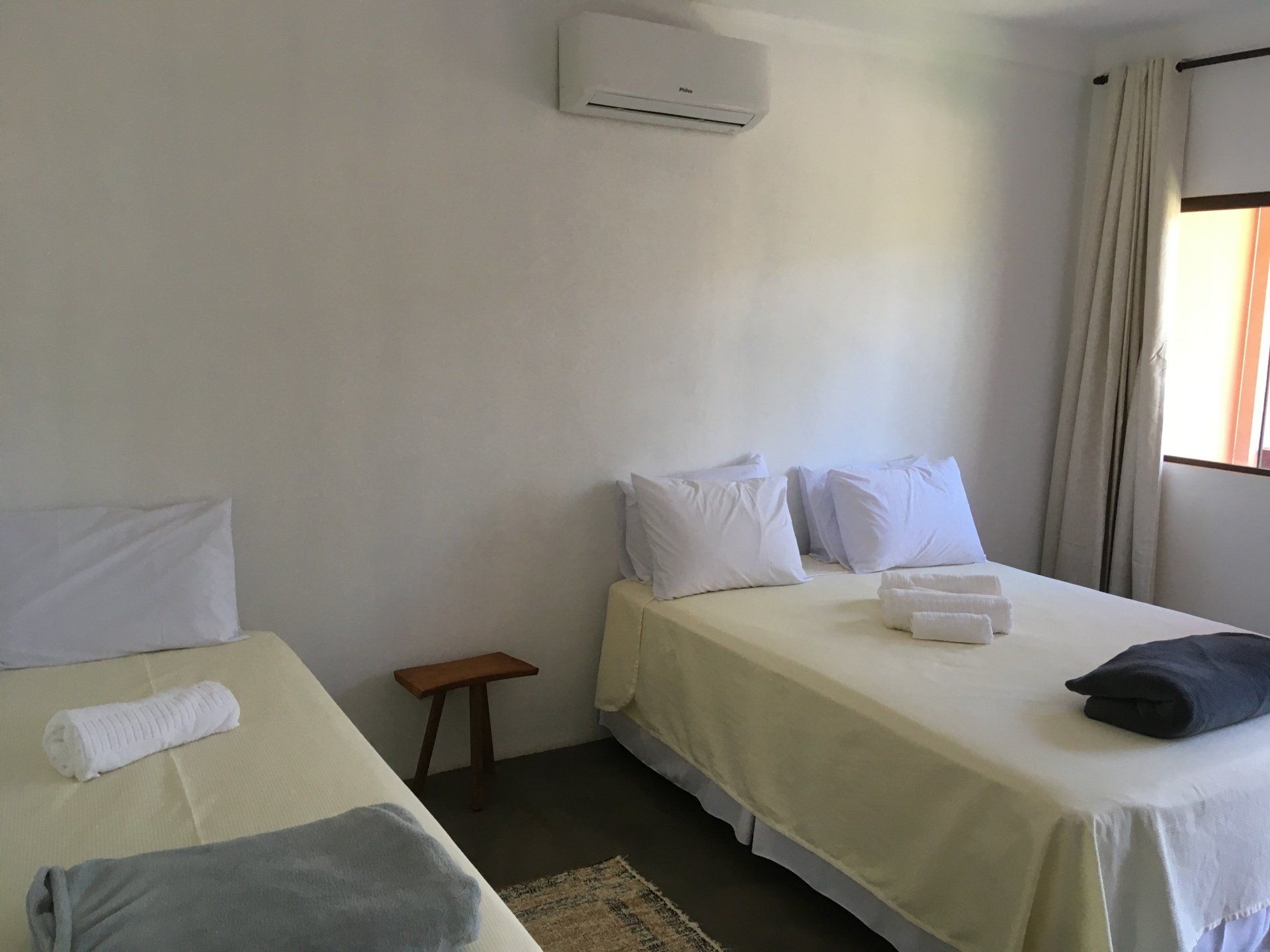 Duas camas de solteiro em um quarto com paredes brancas, um pequeno banquinho de madeira e janela com cortinas.