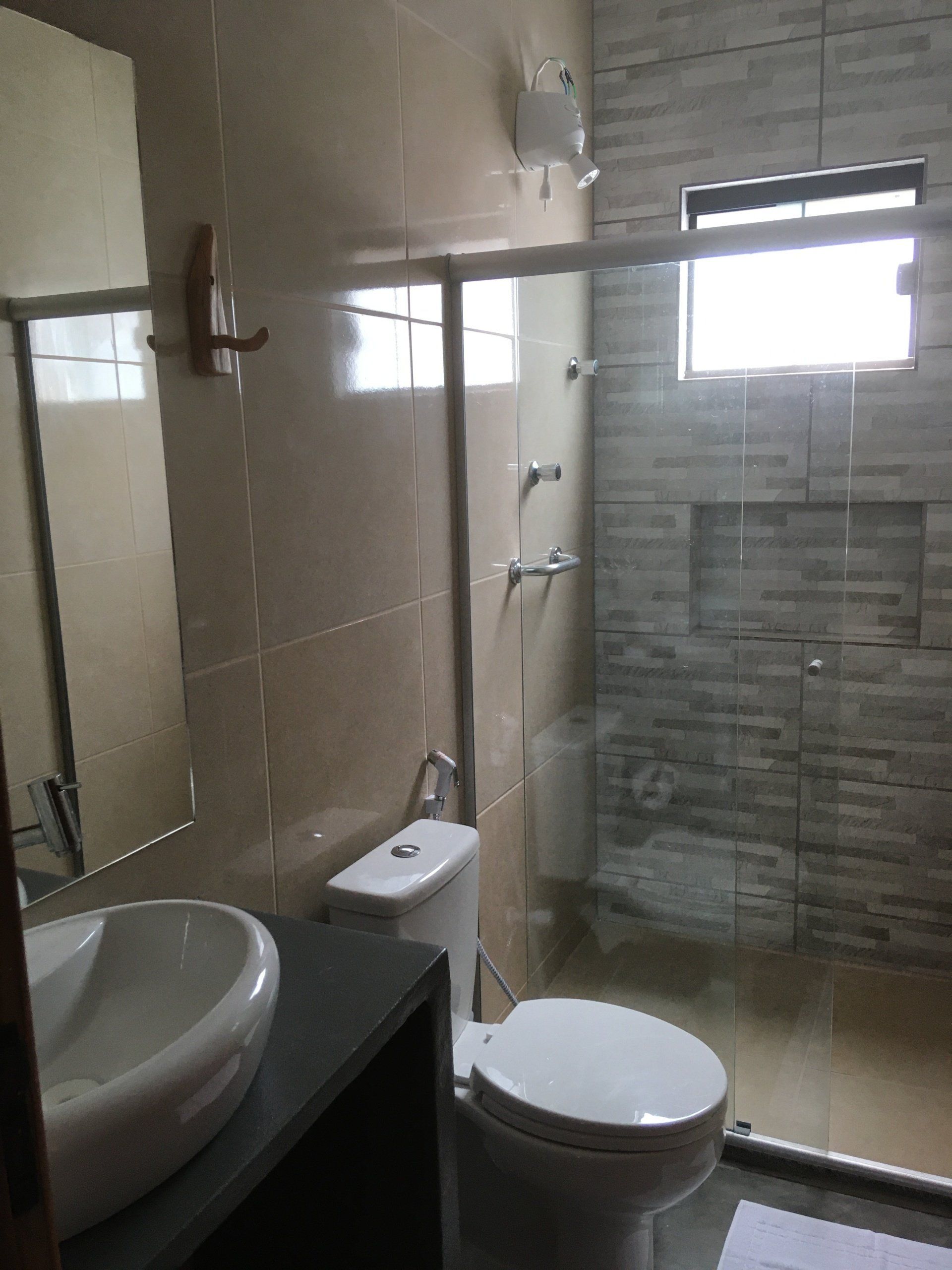 Banheiro com vaso sanitário, pia, chuveiro de vidro e janela pequena; paredes bege, detalhes em cinza.