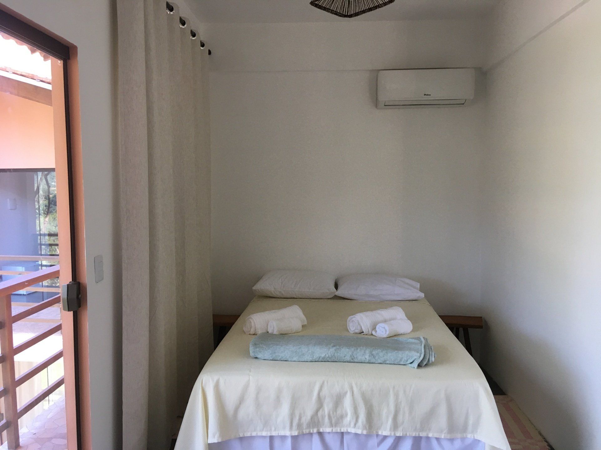 Um quarto pequeno e iluminado com uma cama, cortinas e uma porta aberta para uma varanda.