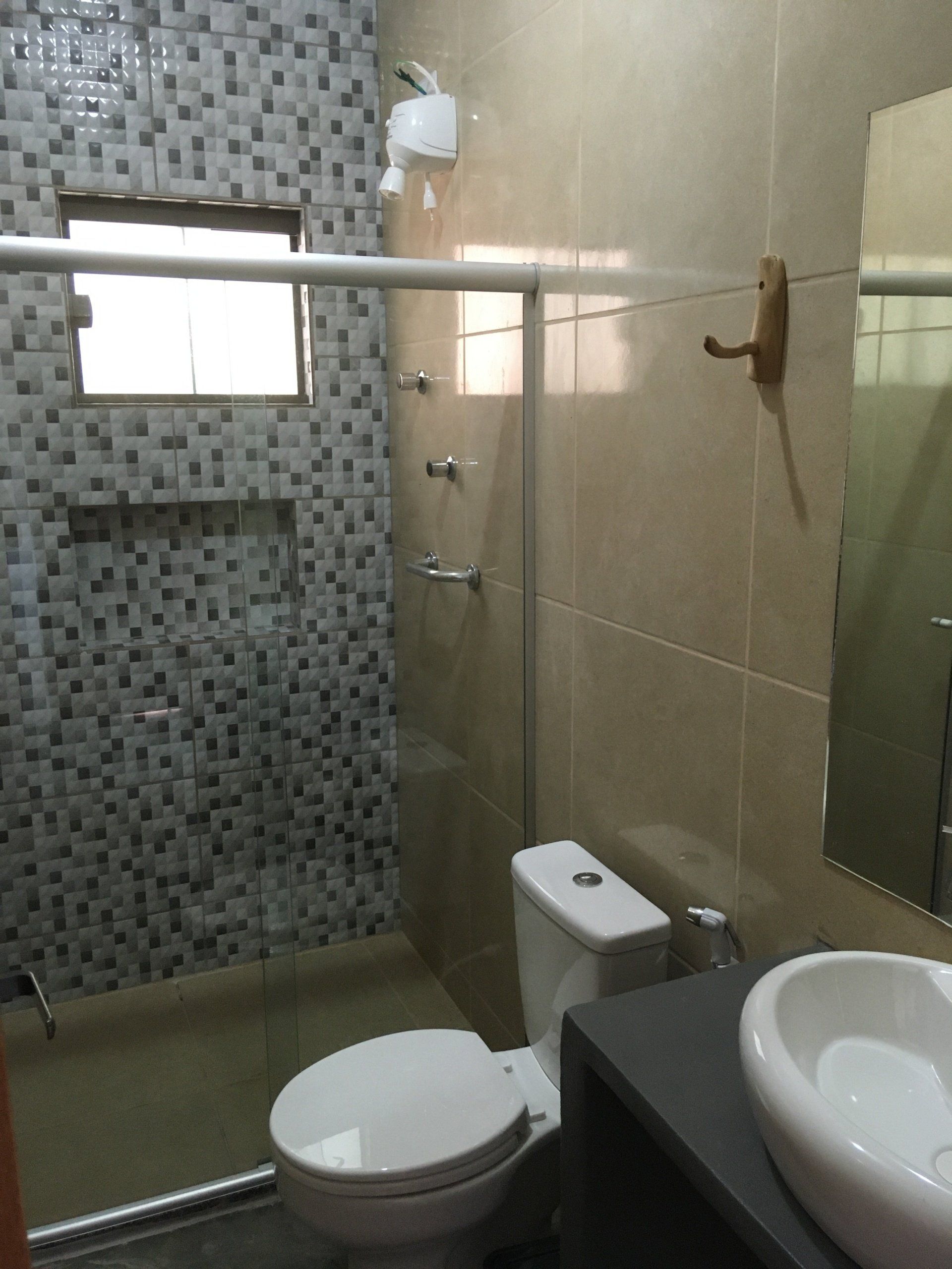 Banheiro com chuveiro, vaso sanitário, pia e porta de vidro revestidos em azulejo. Azulejos e acessórios claros e escuros.