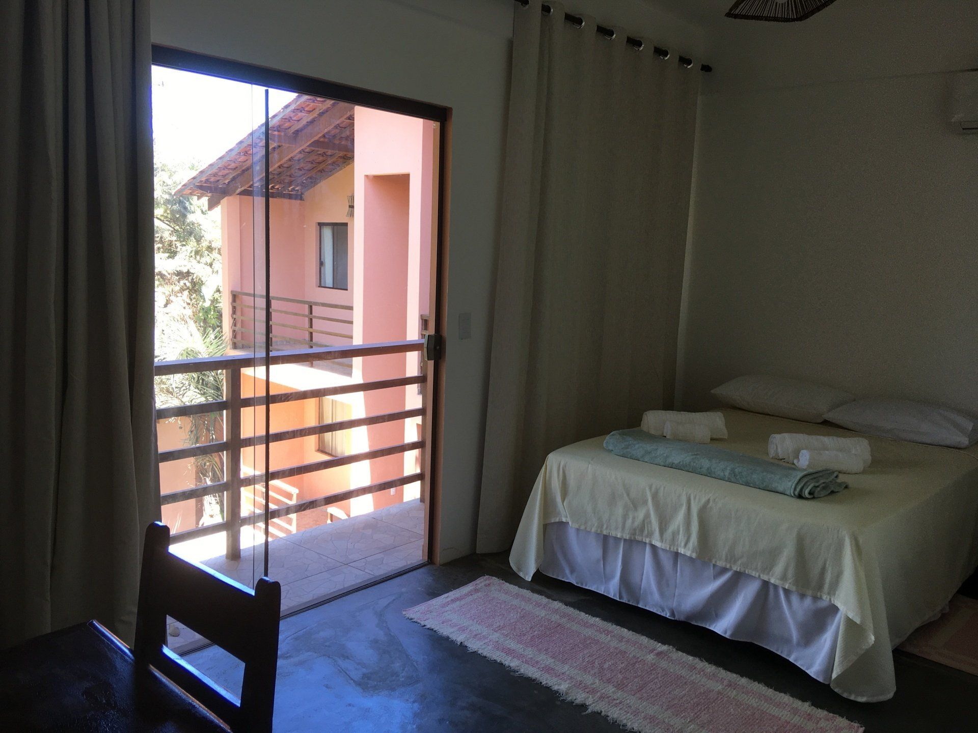 Quarto com cama, vista para a varanda, cortinas bege e um tapete rosa.