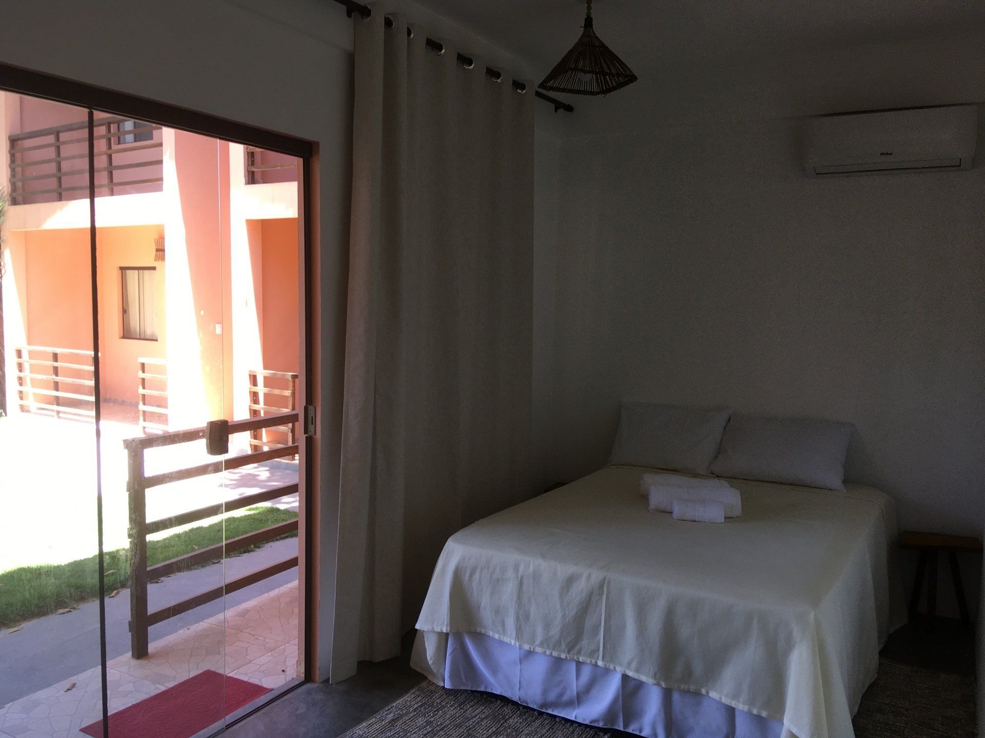 Quarto com cama de casal, porta de vidro para varanda, lençóis brancos, cortinas creme.