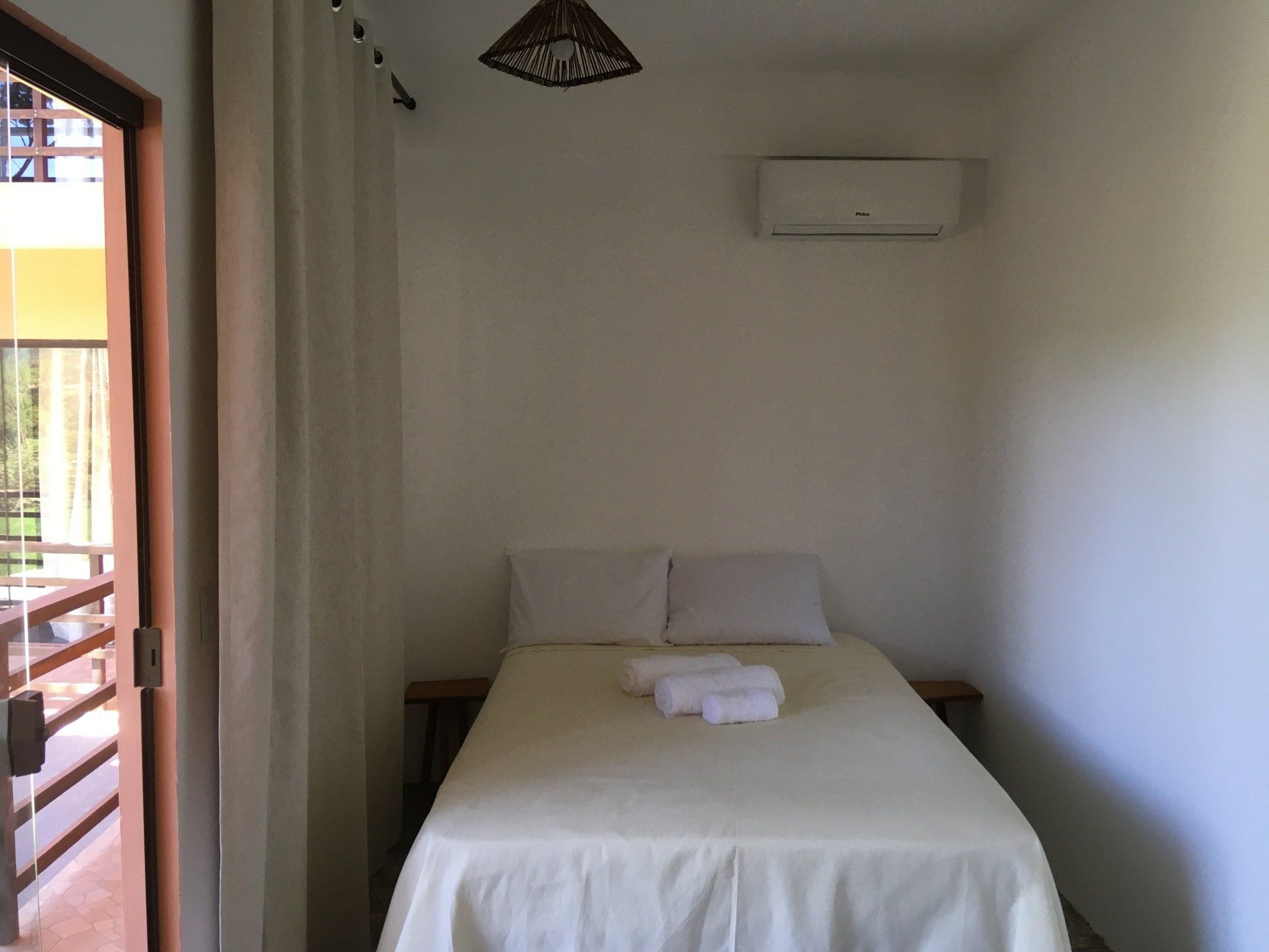 Quarto pequeno com cama, lençóis brancos, travesseiros, toalhas e ar-condicionado. Porta aberta para a varanda.