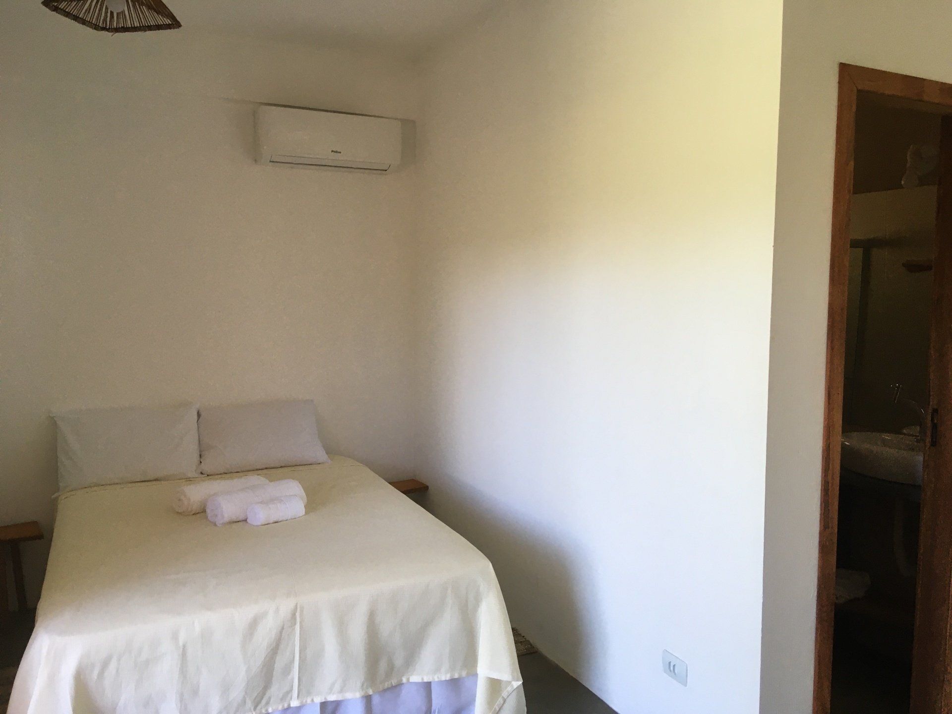 Quarto com cama, toalhas, ar-condicionado e porta para banheiro. Paredes brancas.