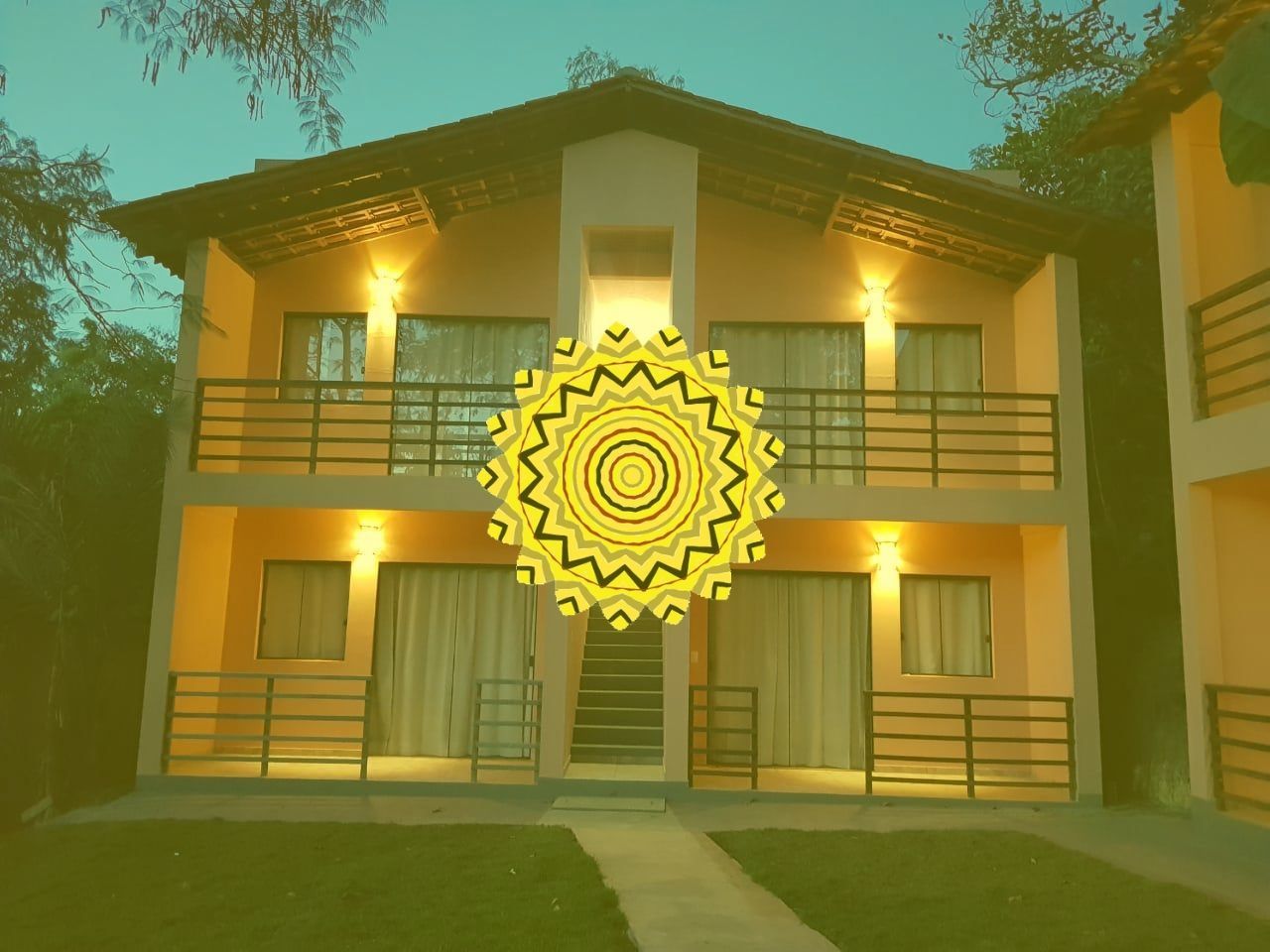 Prédio de dois andares com varandas e luzes acesas, centralizado por um desenho de sol amarelo.