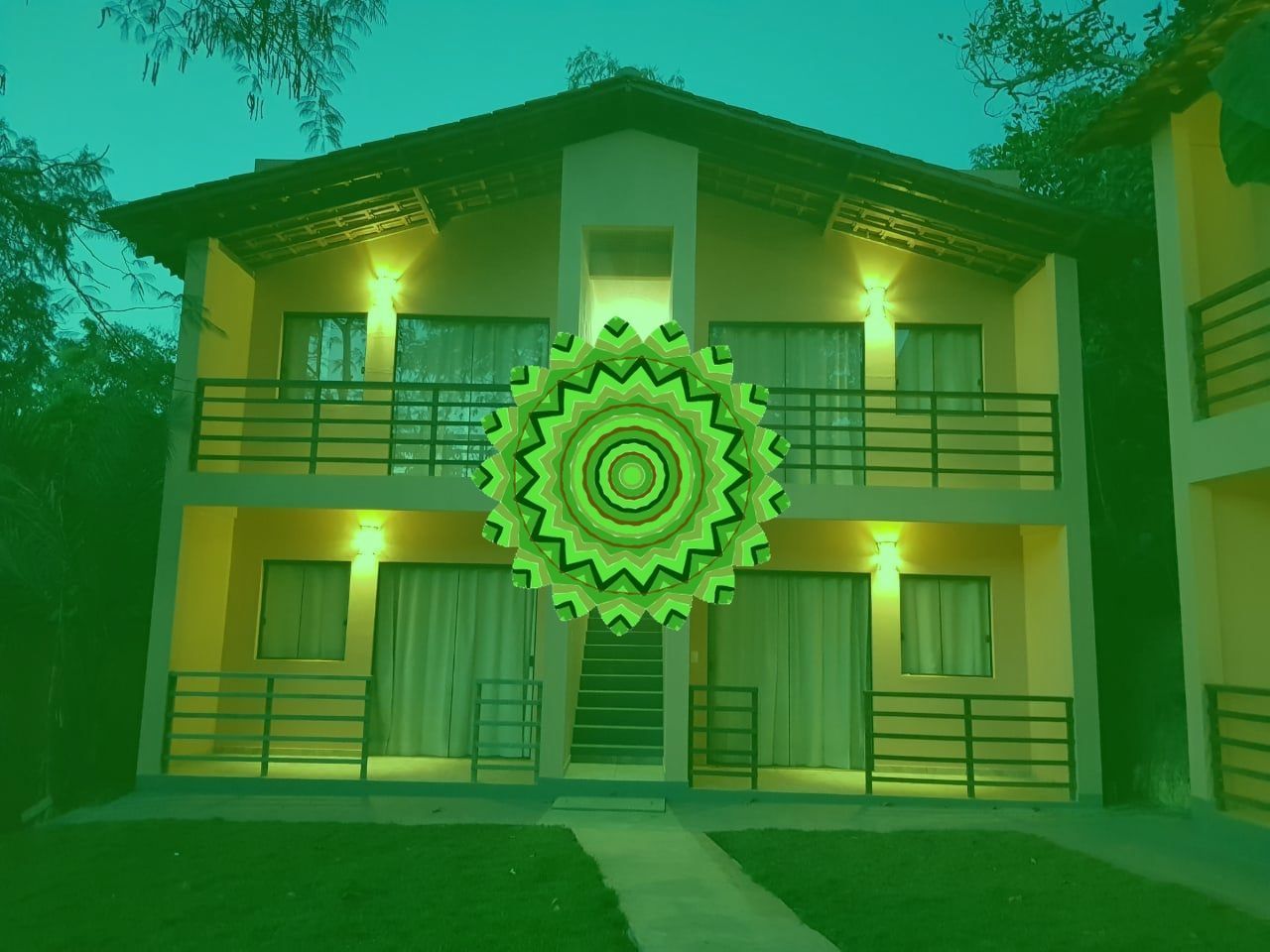 Prédio de dois andares com varandas e cobertura verde, com uma mandala na frente.