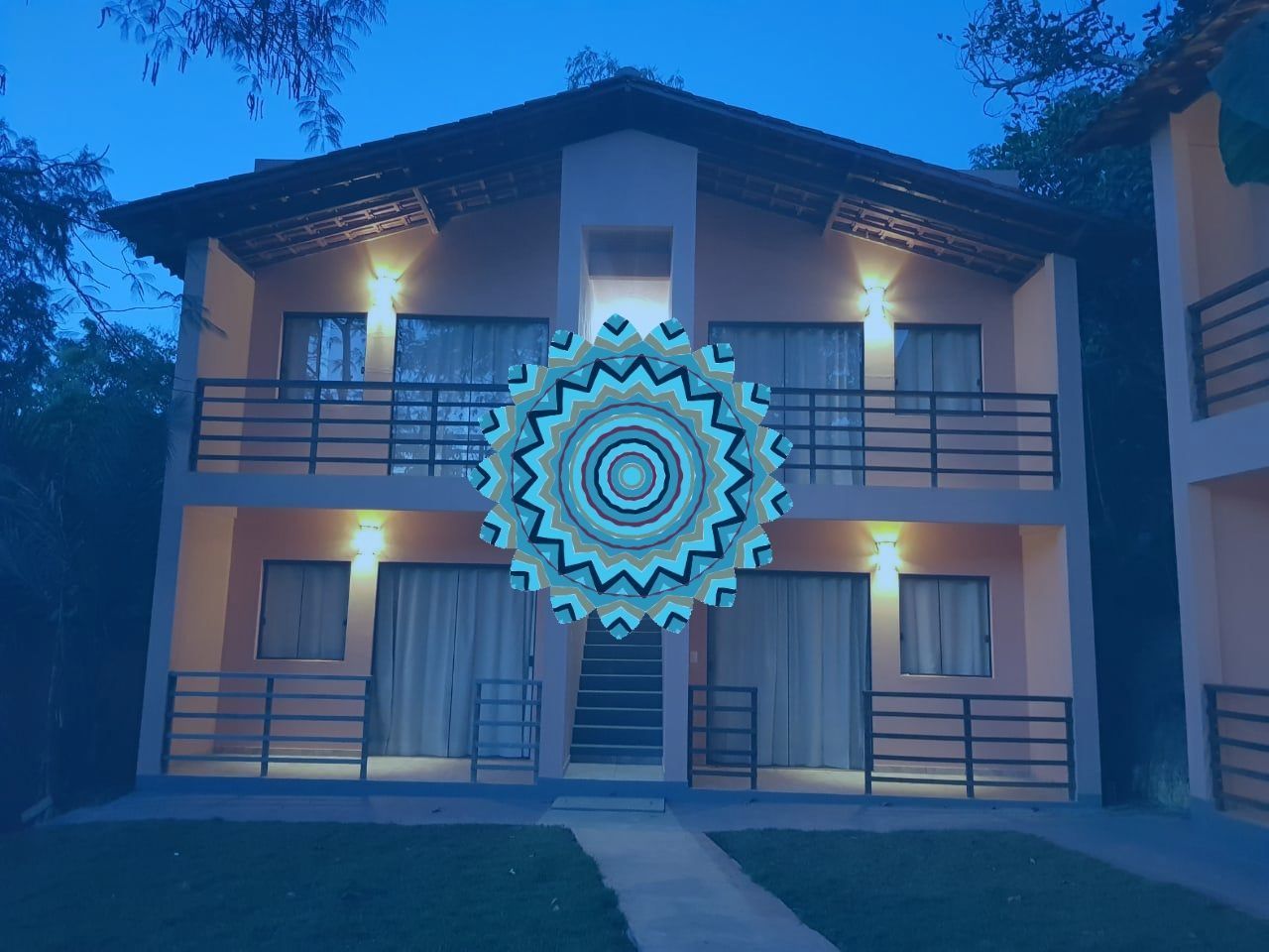 Edifício de dois andares com varandas, iluminado ao anoitecer e com uma mandala brilhante no centro.