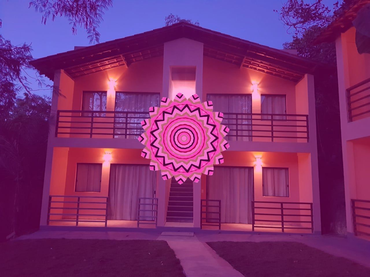 Edifício de dois andares com varandas iluminadas por luzes quentes contra um céu crepuscular; uma mandala rosa sobreposta no centro.