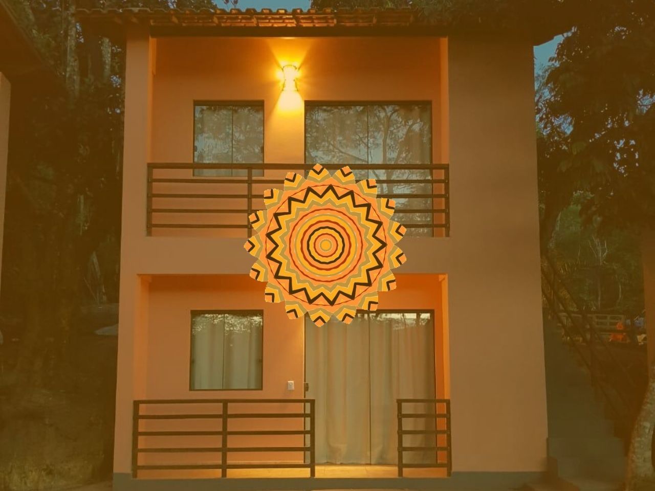 Prédio de dois andares na cor pêssego, com varandas; mandala dourada sobreposta. Floresta ao fundo.