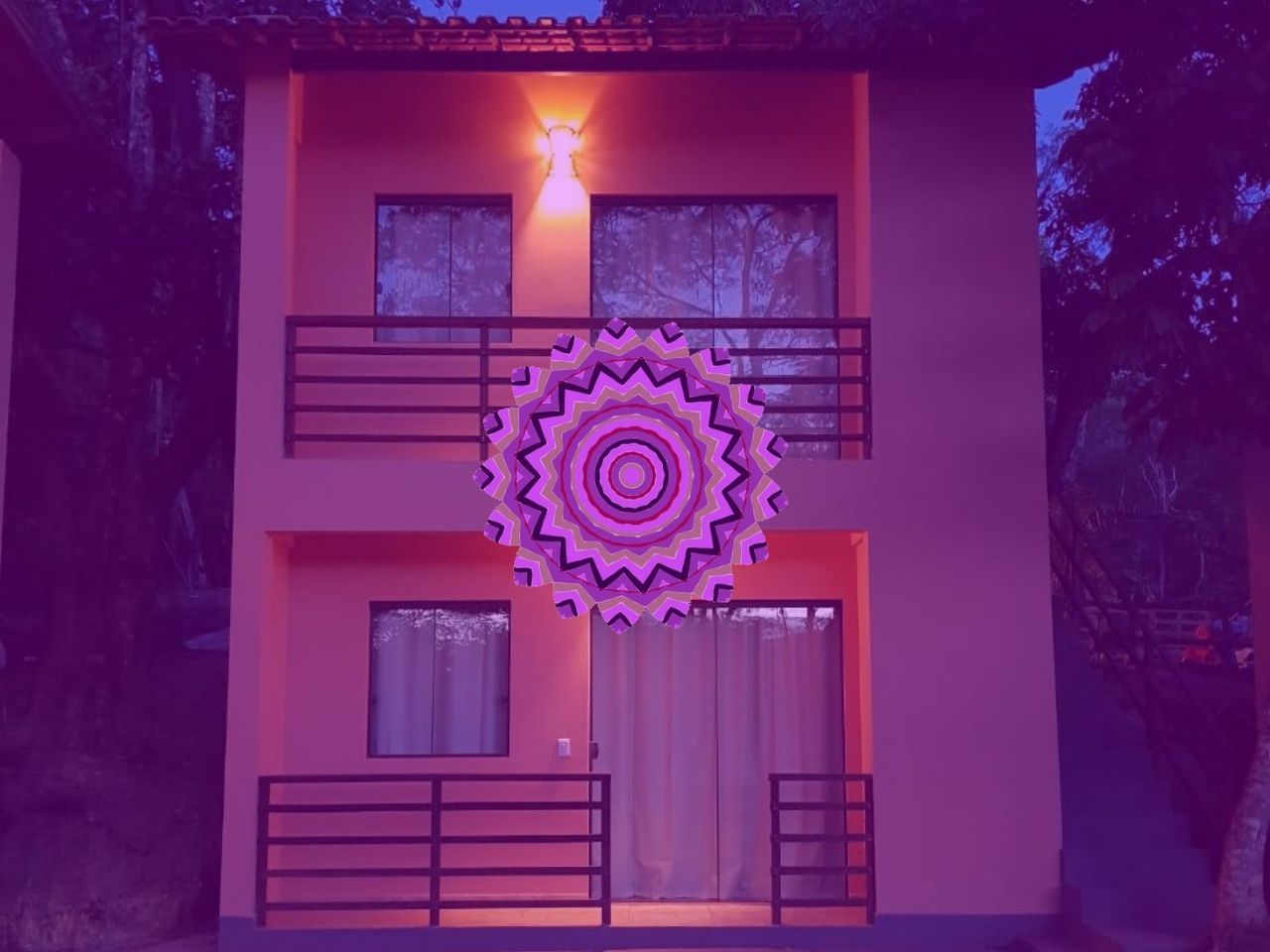 Casa de dois andares na cor pêssego, com grades pretas, iluminada por uma mandala rosa-púrpura.