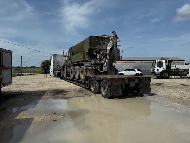 A military vehicle is being transported on a trailer in a muddy parking lot.