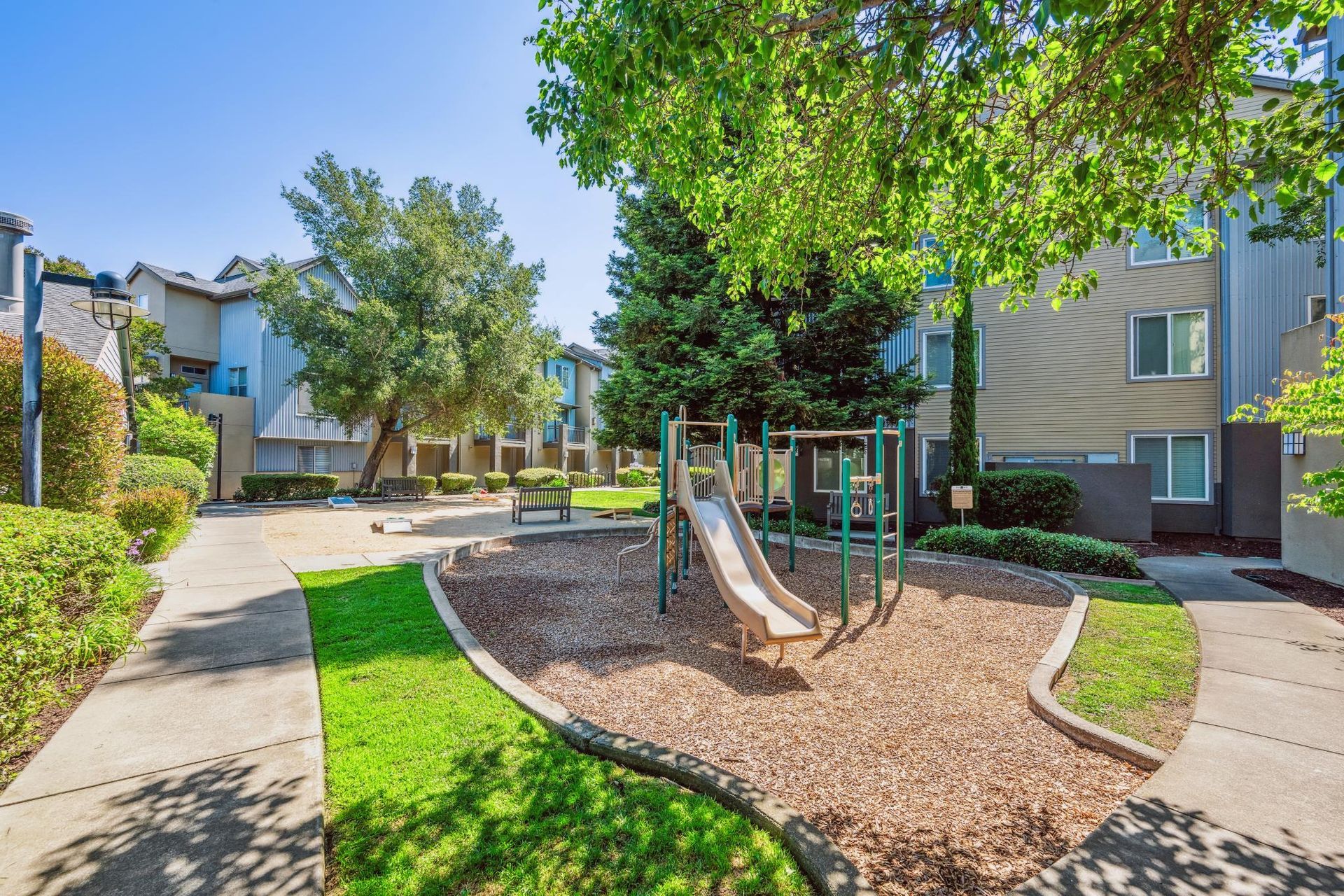 Playground with slide surrounded by wood chips, grass, and apartment buildings.