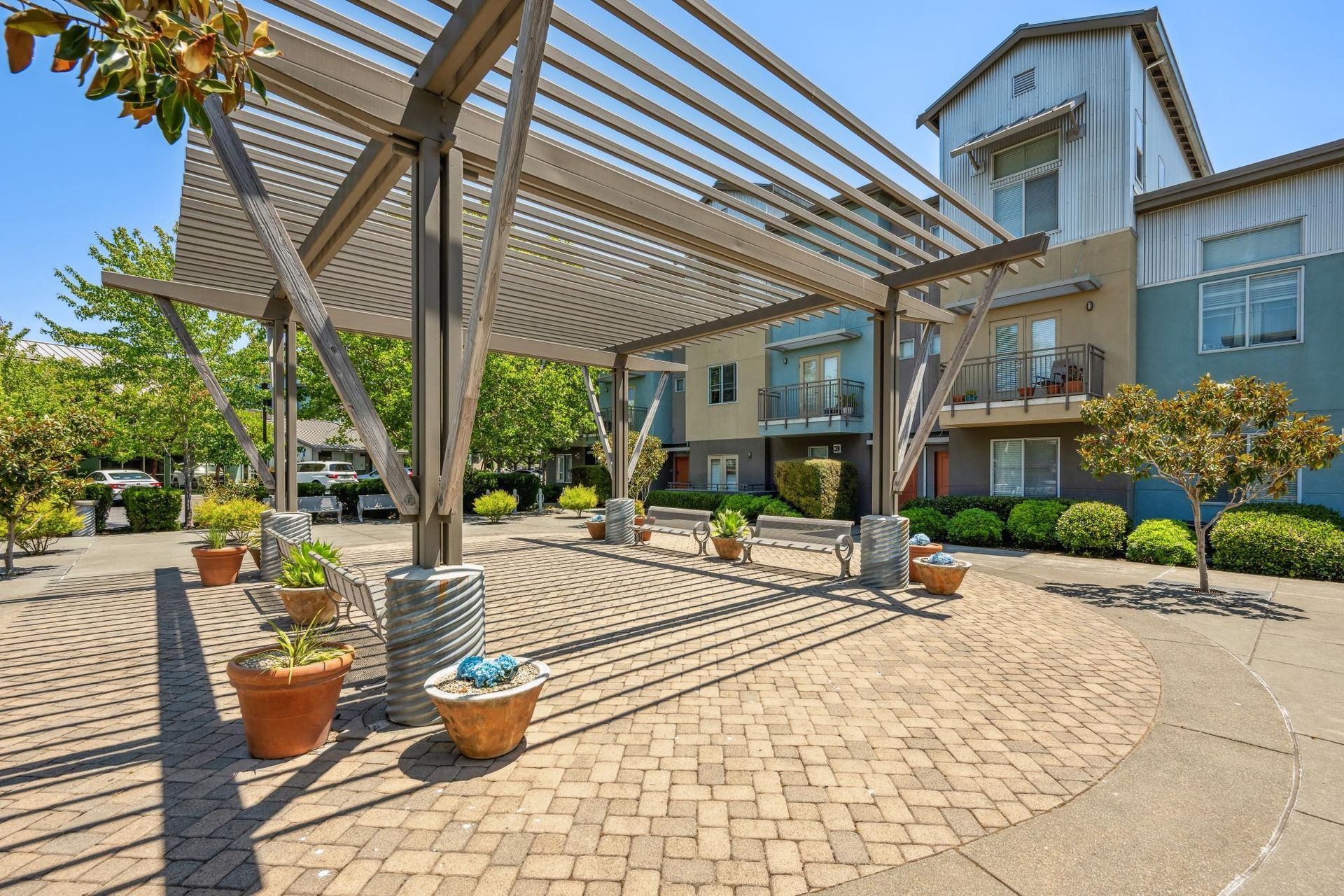 Outdoor patio with pergola, brick pavers, potted plants, and colorful buildings in background.