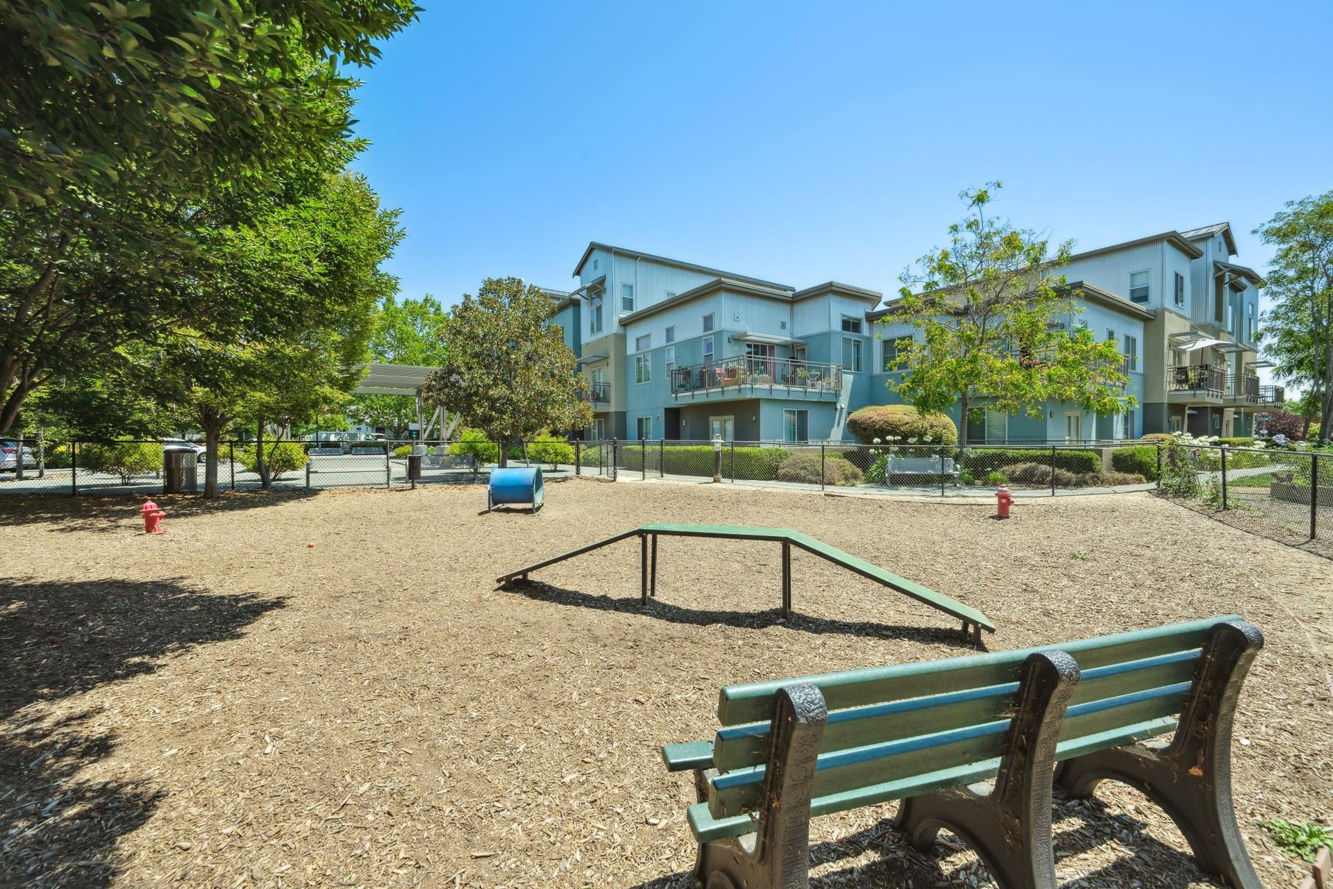 Dog park with agility equipment, wood chip ground cover, benches, and apartment buildings in the background.