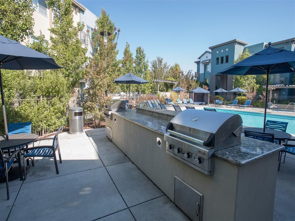 Outdoor community pool area with grill, tables, umbrellas, and lounge chairs.