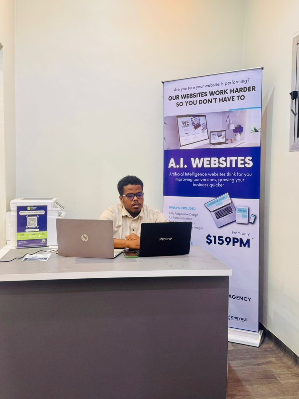 Man sits at a desk with two laptops, in front of a banner promoting AI websites for $159/month.