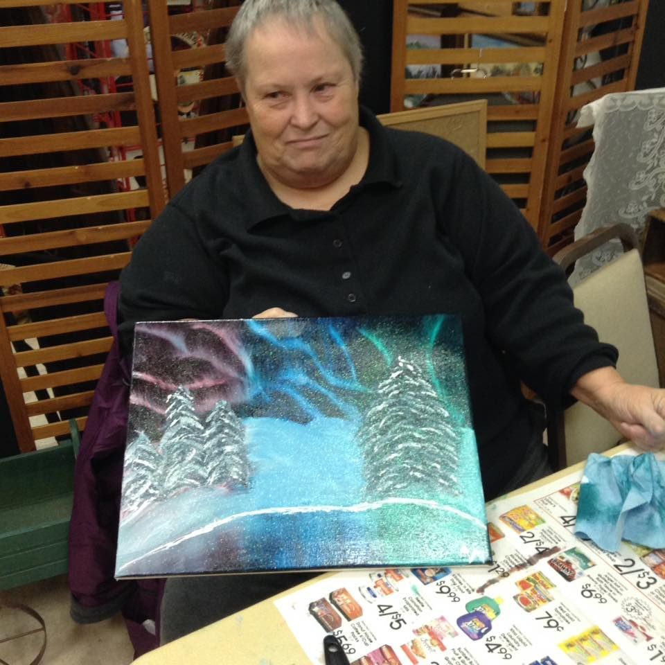 A woman is sitting at a table holding a painting of snowy trees