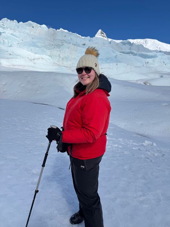 A woman in a red jacket is standing in the snow holding hiking poles.