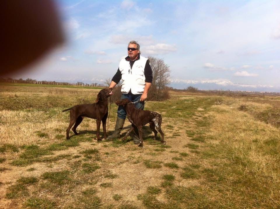 Uomo in un campo con due cani marroni, con indosso un gilet bianco. Giornata di sole, erba secca.