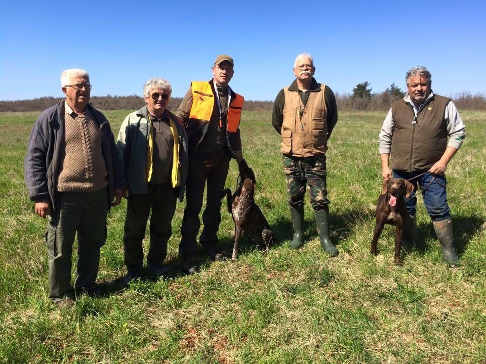 Cinque persone e due cani in un campo, probabilmente a caccia. Gli uomini indossano gilet, stivali e tengono il guinzaglio. Giornata di sole.