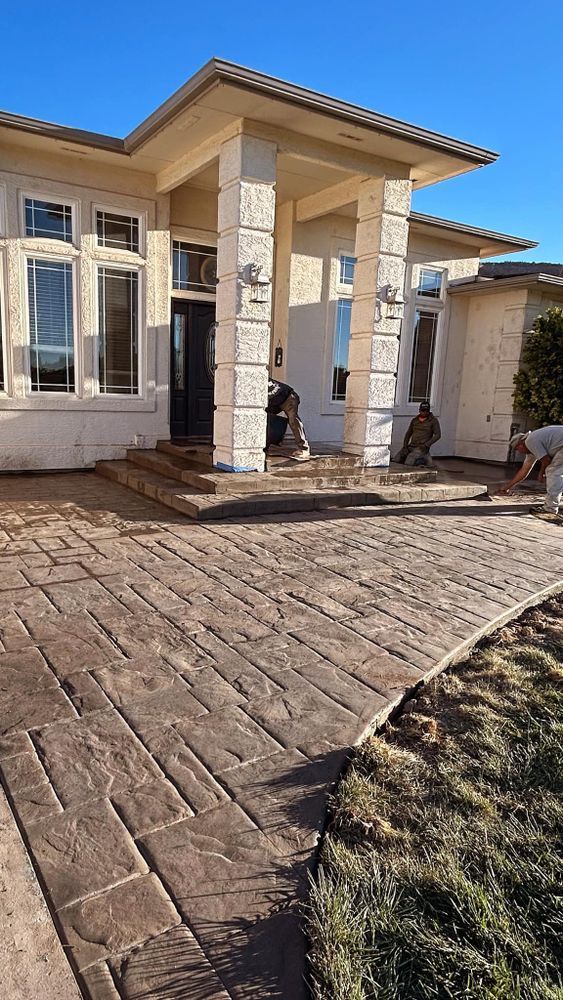 Brick-patterned concrete walkway leading to a house with columns. People work near the entrance on a sunny day.