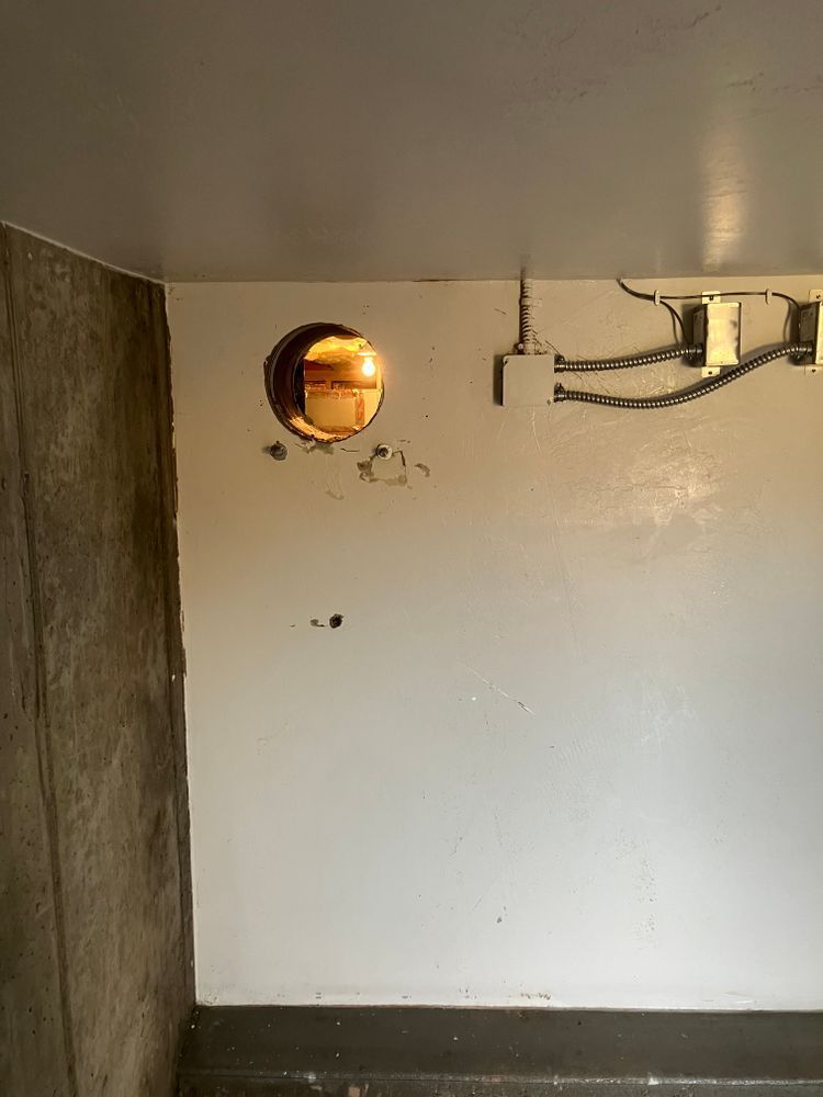 Hole in a white wall; electrical wires, a concrete wall to the left, and a metal beam above.