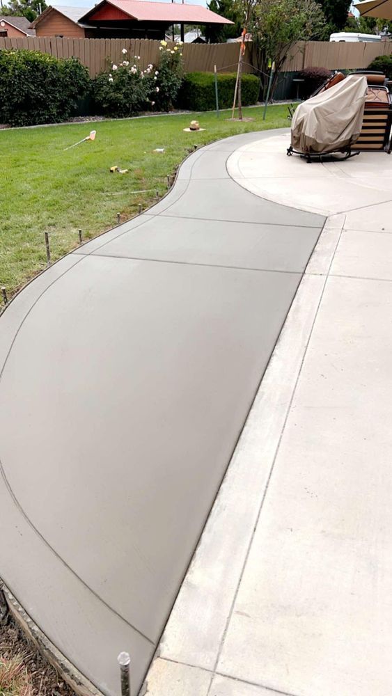 Freshly poured concrete walkway curving through a backyard, connecting to an existing patio.