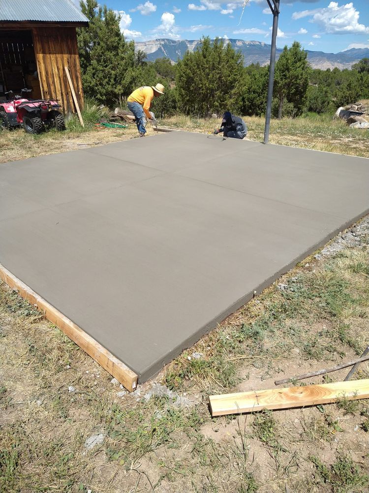 Two people smoothing a newly poured concrete pad in a rural setting, with a wood shed in the background.