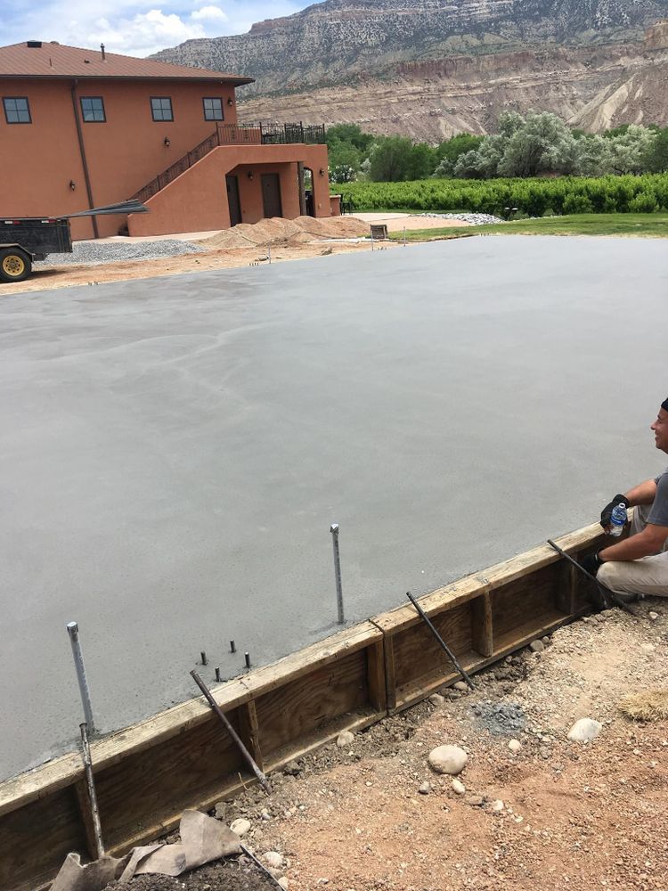 Newly poured concrete slab with a man working near a wooden border, in front of a building and mountain.