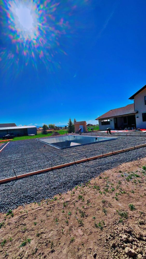 Construction site with pool foundation, gravel, and house under bright blue sky.