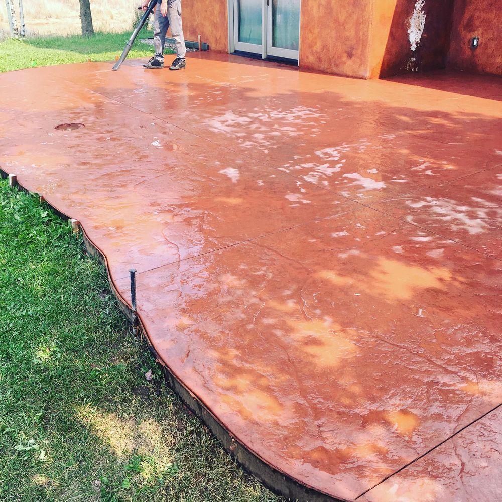 Man pressure washing a red-stained concrete patio near green grass and a house.