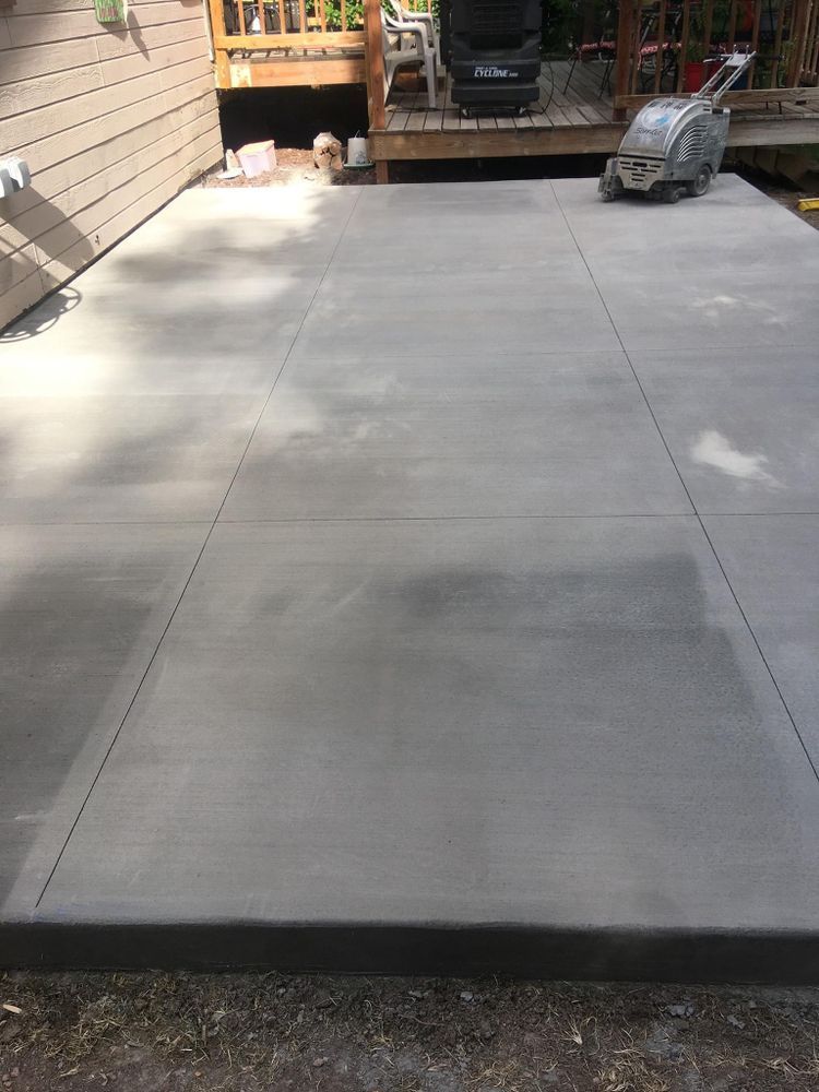 Newly poured concrete patio, gray color, with control joints, near a wooden deck and siding.