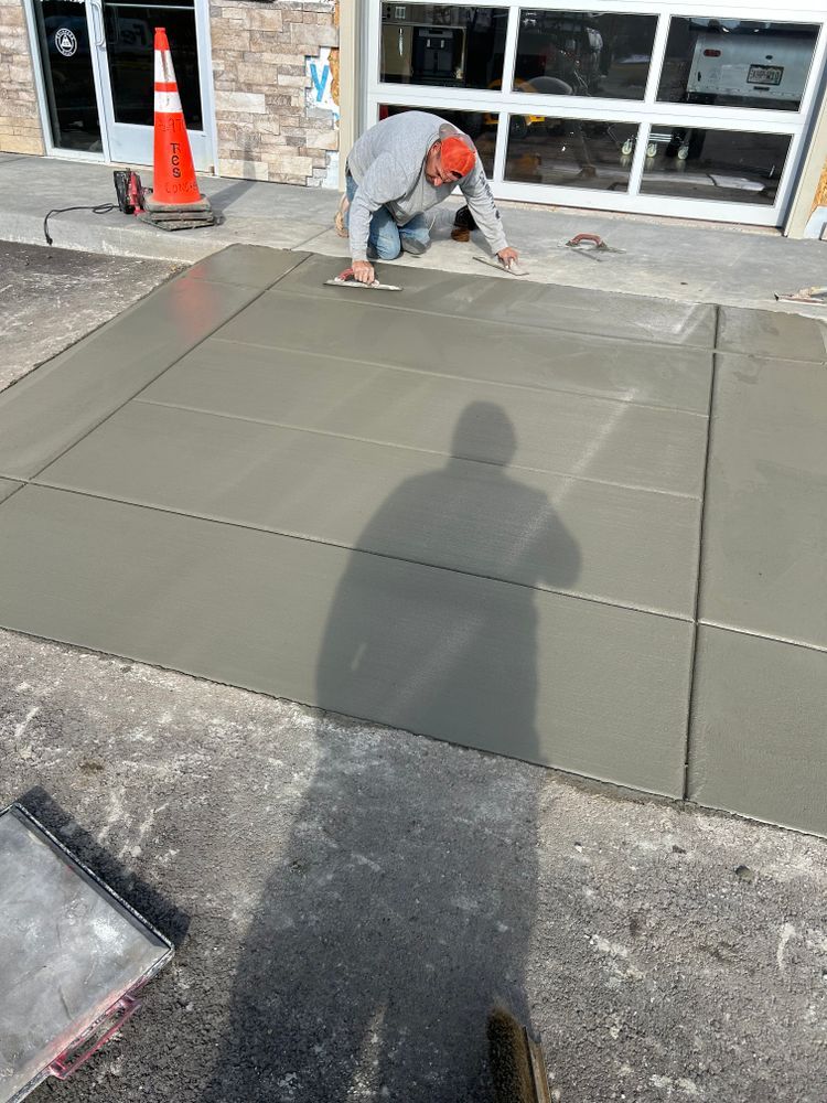 Man troweling wet concrete sidewalk in front of a building with a garage door and an open door.