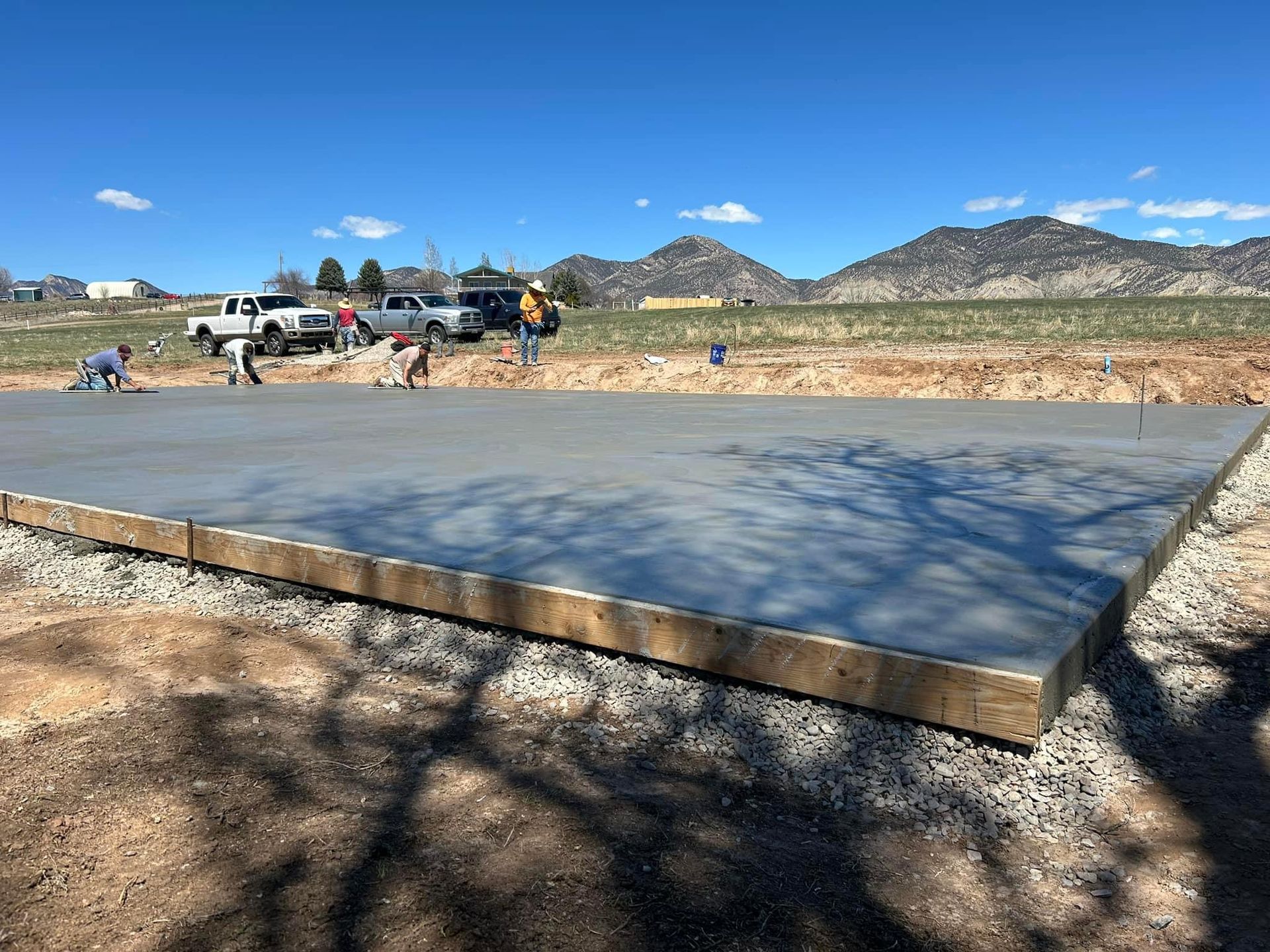 Workers smoothing a fresh concrete slab outdoors, with trucks and mountains in the background under a blue sky.