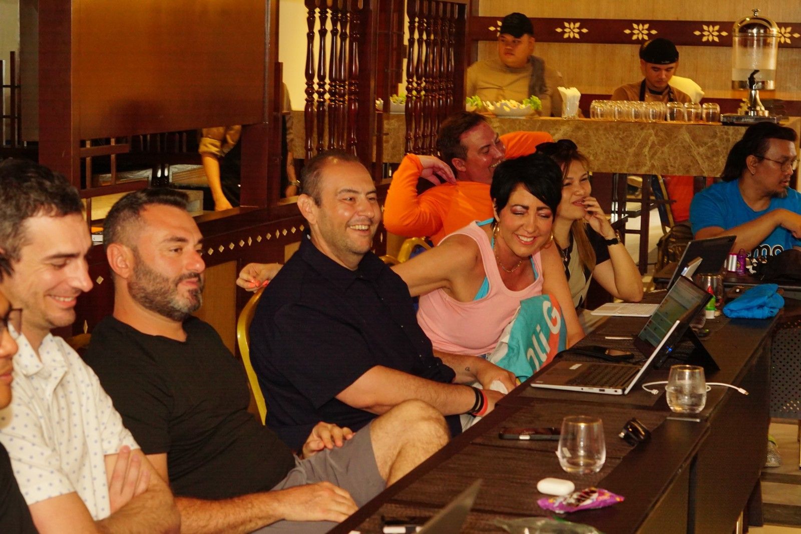 People seated at a table, looking at laptops and smiling. Indoors, some people in background.