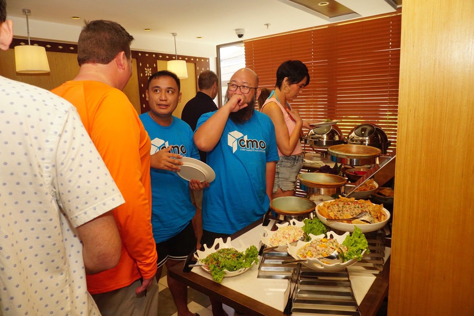 People at buffet: Man in blue shirt tastes food, others select dishes.