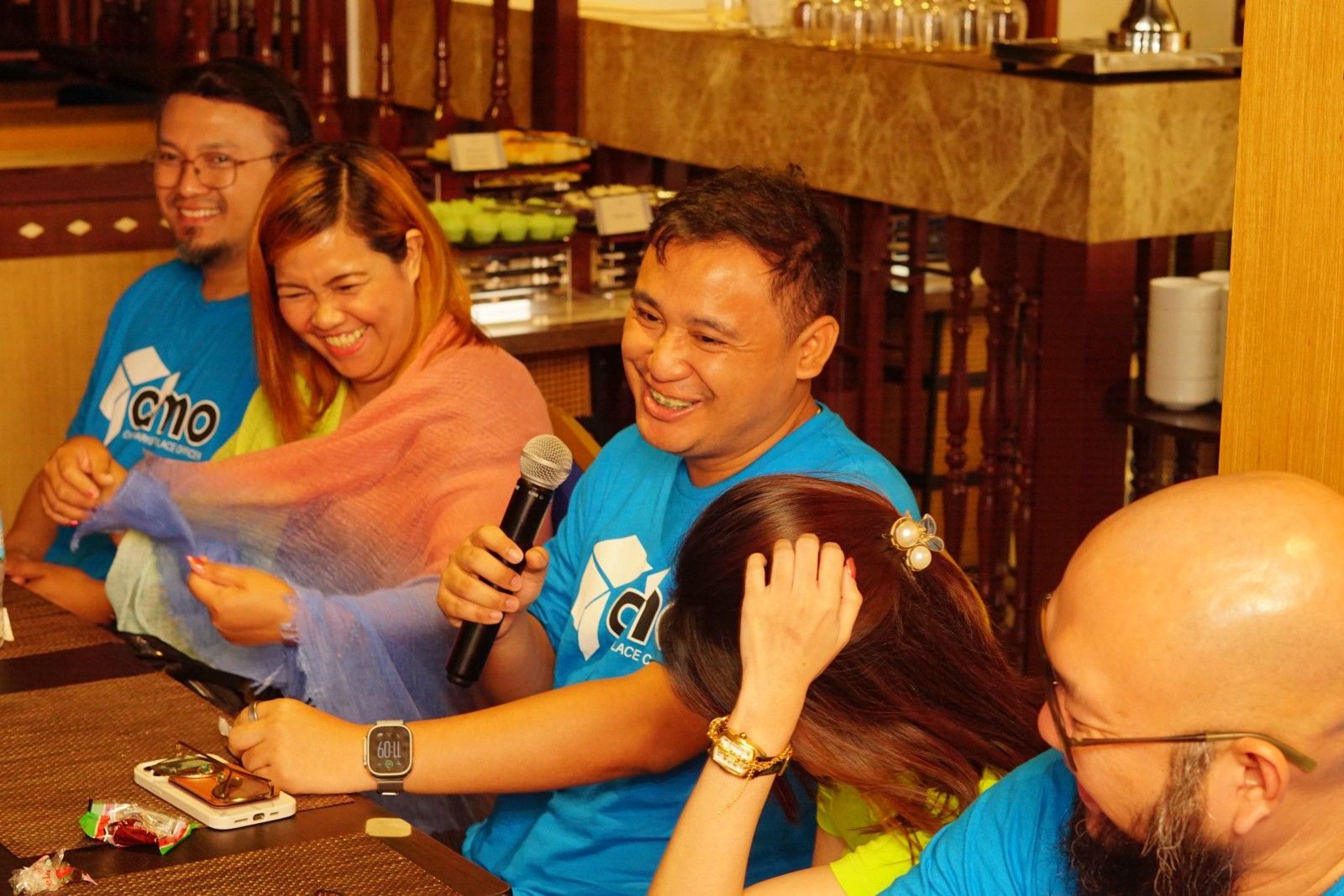 People at a table, some smiling and laughing. One person holds a microphone.