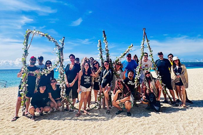 Group of people posing by a beach with a decorative floral arch spelling 