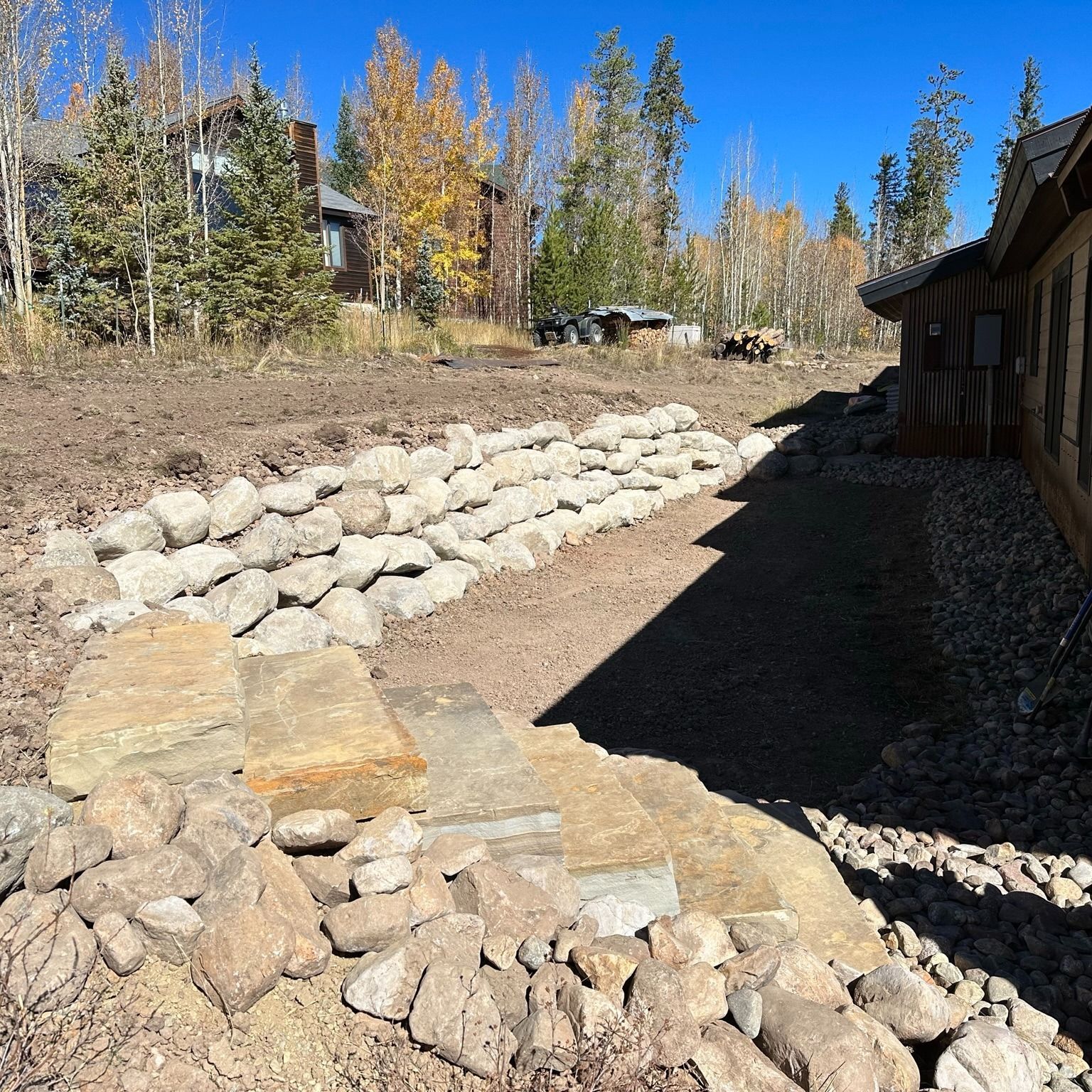 Stone retaining wall with steps, next to a building and dirt area, blue sky background.