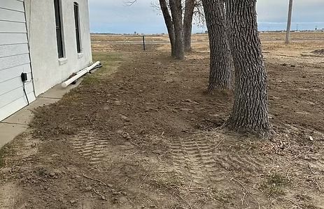 Side of a white building with bare dirt, trees, and cloudy sky. Tire tracks visible.
