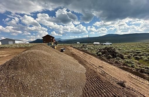 Gravel pile on dirt road, a small building in the background. Cloudy sky, mountains in the distance.