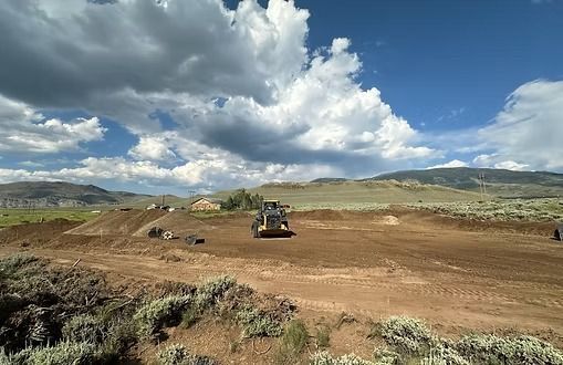 A yellow bulldozer levels dirt in an open field under a blue sky with fluffy clouds.